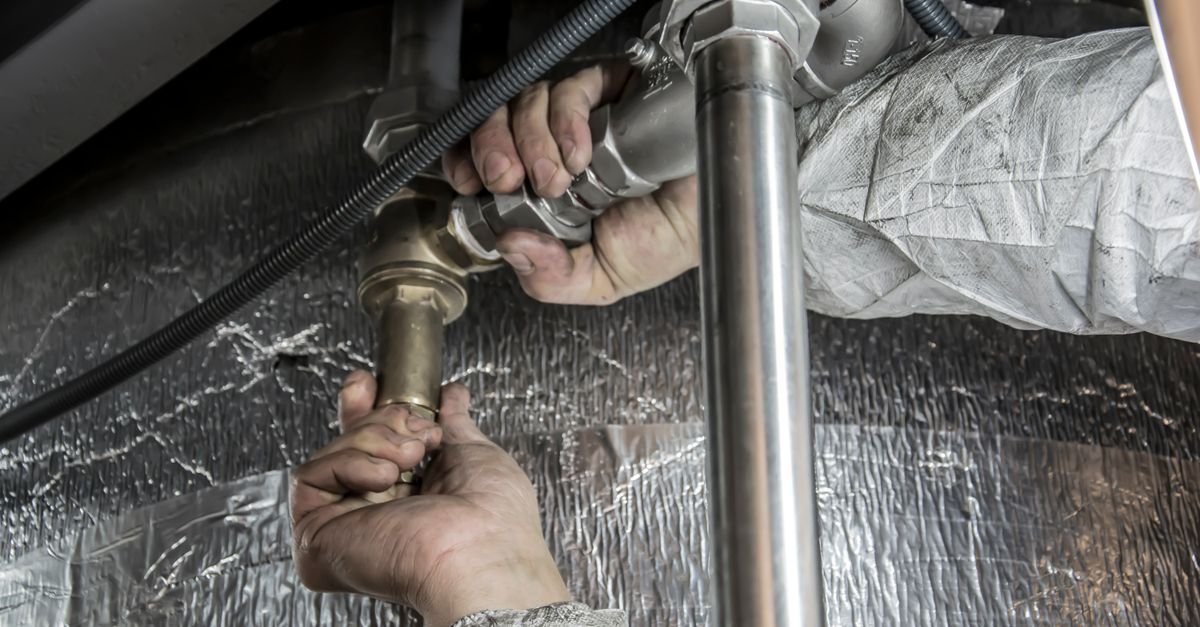 Close-up of a plumber's hands fitting steel pipework, showing the inline connections a chemical injector sits between