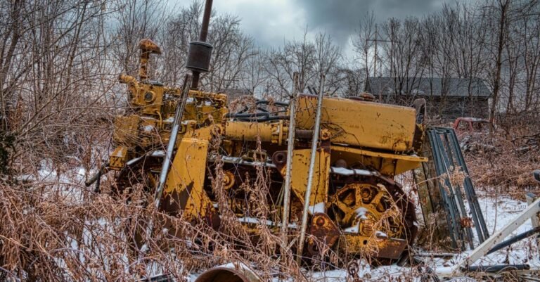An abandoned rusty tractor sitting in deep snow among bare winter trees, the way equipment gets left out over a cold season