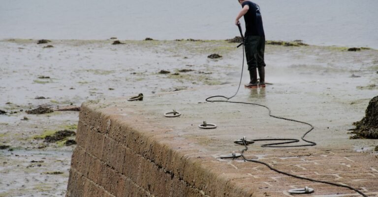 Worker gripping a pressure washer gun with both hands while cleaning a seaweed-covered stone pier at low tide