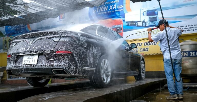 A man gripping a pressure washer wand outdoors, watching the spray pattern as he rinses a black car
