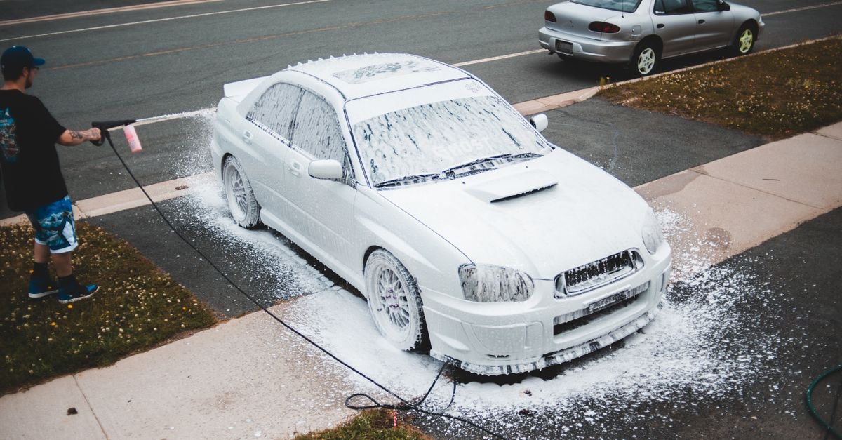 A man rinsing a foam-covered car from above with a pressure washer gun, the wide low-pressure soap tip still fitted