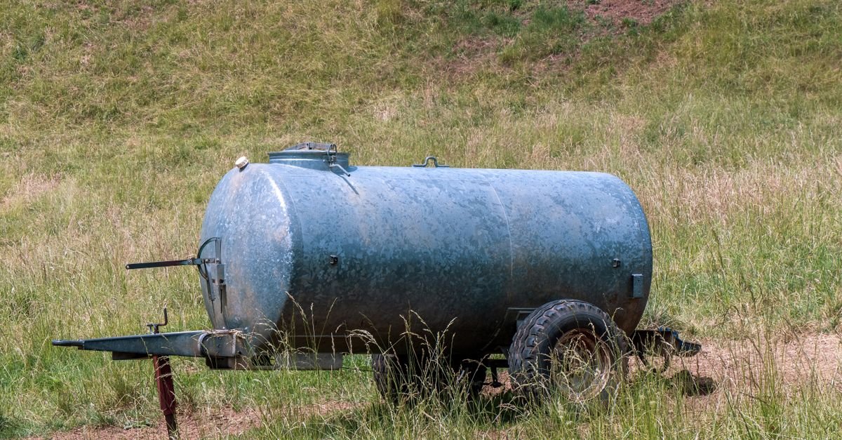 A metal water tanker standing in a field, the sort of gravity supply that offers no pressure to hold a hose open