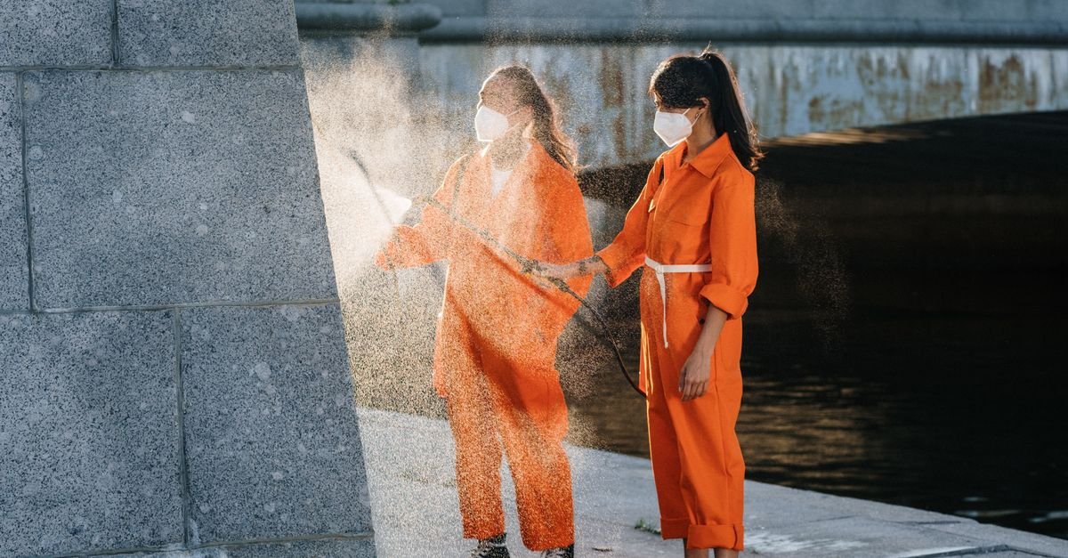 Two workers in orange jumpsuits cleaning an outdoor surface with water in daylight