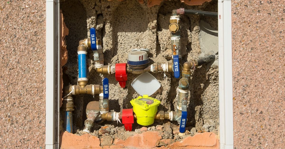 A complex domestic water meter assembly set into a concrete wall, the supply-side plumbing that decides how many gallons per minute ever reach a pressure washer