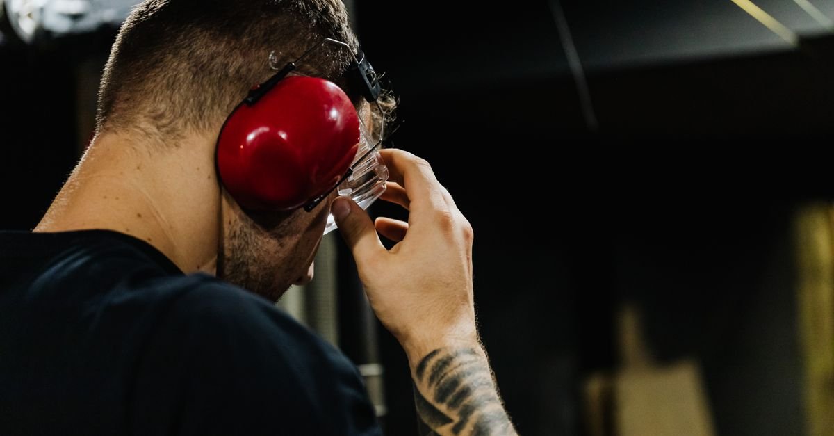 A man in a workshop fitting red over-ear defenders alongside safety glasses, the protection a gas pressure washer's noise level actually calls for