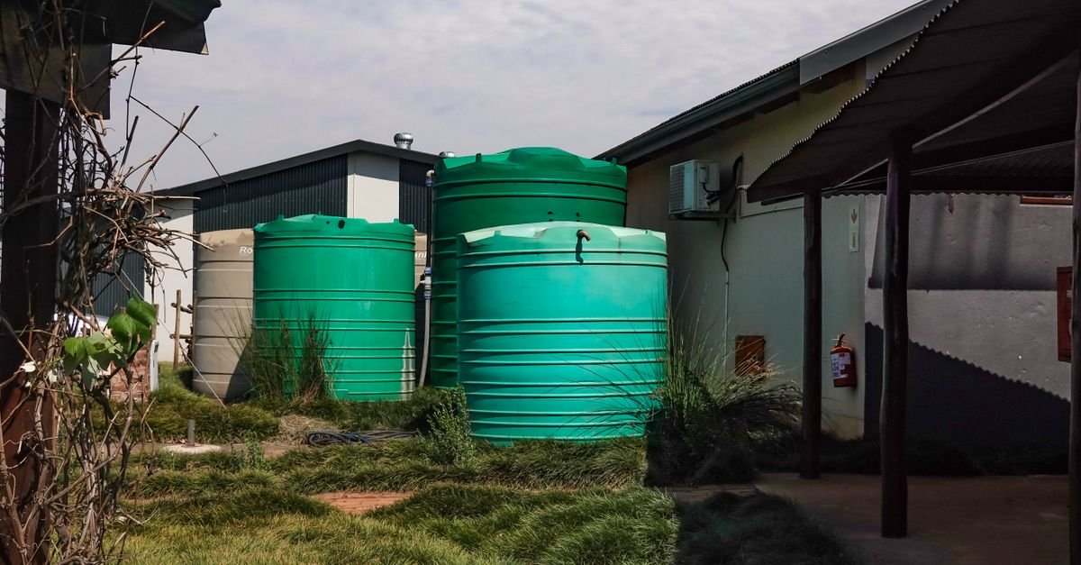 Three green water storage tanks standing beside a rural farmhouse, the sort of buffer supply that lets a slow tap feed a fast pump