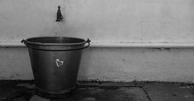 An aged metal bucket standing under an outdoor faucet against a rustic wall, the setup for timing a water supply flow rate