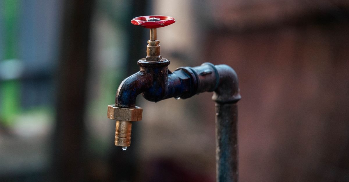 A single water droplet falls from the spout of a weathered outdoor tap