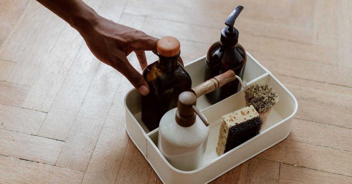Hand reaching for a row of detergent bottles and cleaning brushes on a wooden floor
