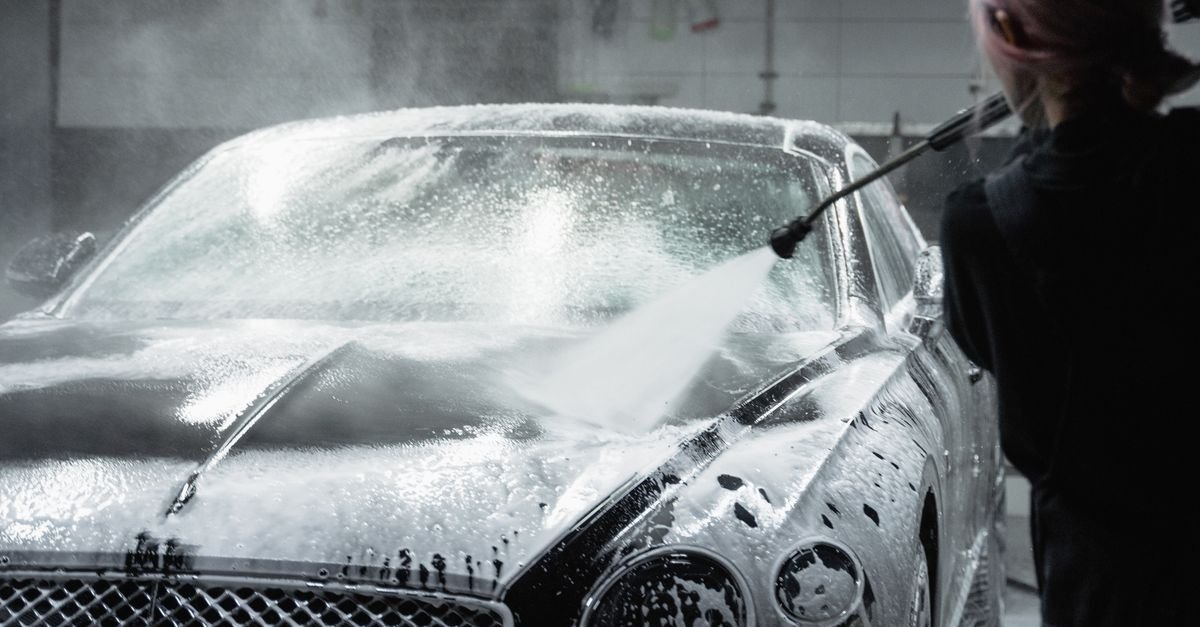 Worker rinsing a dark car coated in white detergent foam inside a garage wash bay