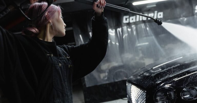 A person aiming a pressure washer lance at a car in a dimly lit garage, where the shape of the spray is easy to see against the paint