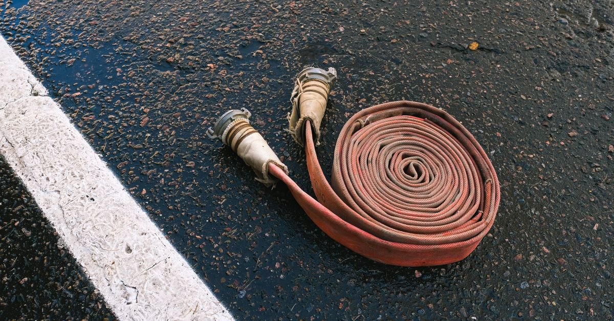 A rolled canvas-jacketed fire hose lying on asphalt, the flexible reinforced wall that gives under a pressure surge instead of passing it on