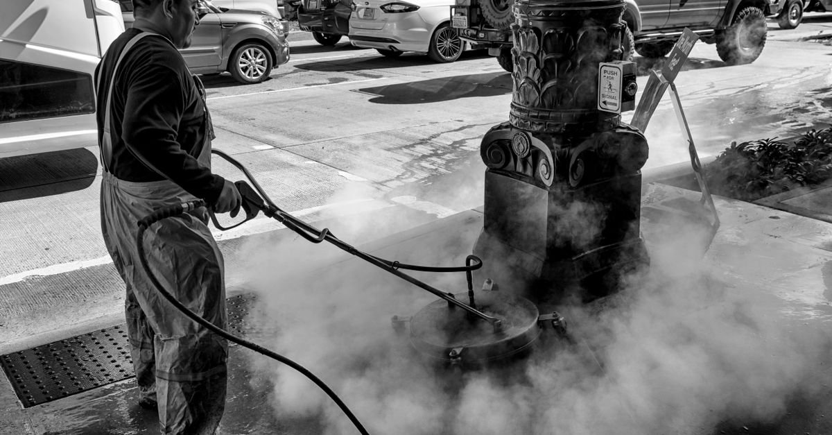 A worker directing a running pressure washer at a fixed street fitting, the machine held at load for minutes at a time