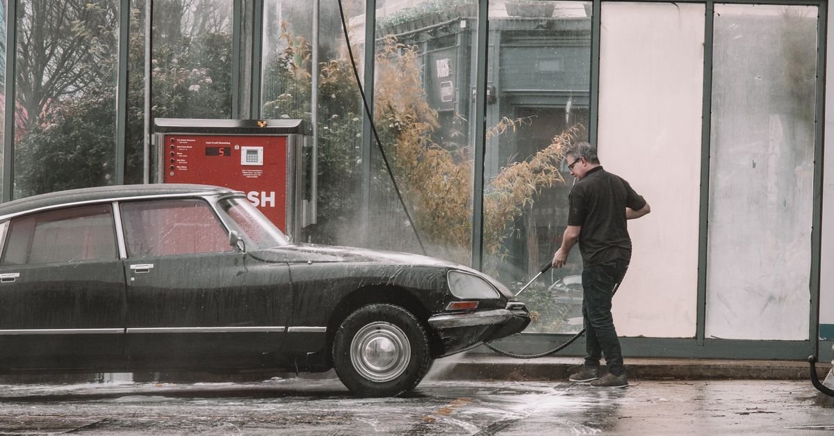 Person using a pressure washer lance to rinse a classic car at an outdoor self-service wash bay