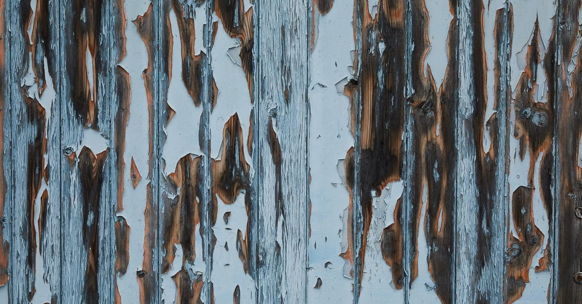 A weathered timber house wall with white paint flaking away from damaged planks