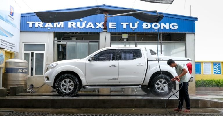 A man cleaning a white pickup truck with a pressure washer at an outdoor wash bay