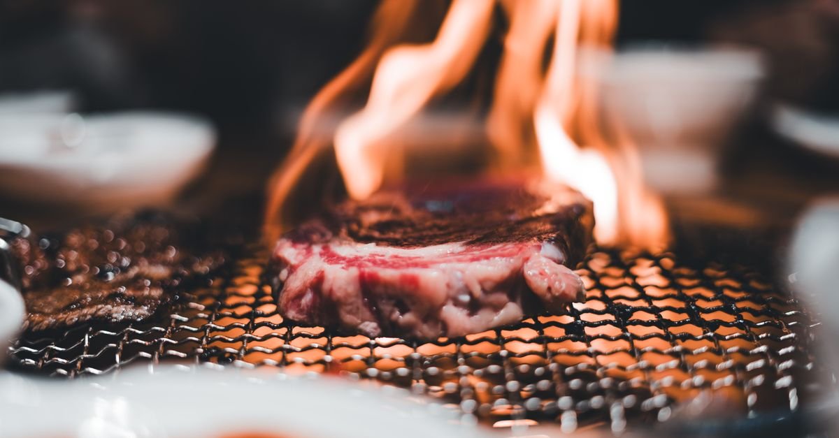 Close-up of a steak on a charcoal barbecue with flames rising from fat dripping through the bars