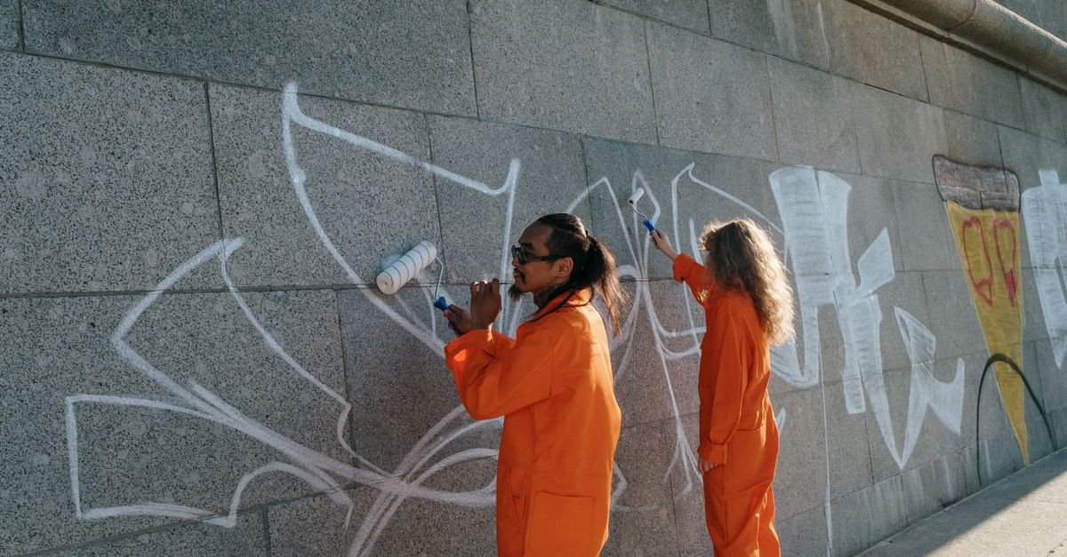 Two workers in orange high-visibility uniforms cleaning spray-painted graffiti from a concrete wall