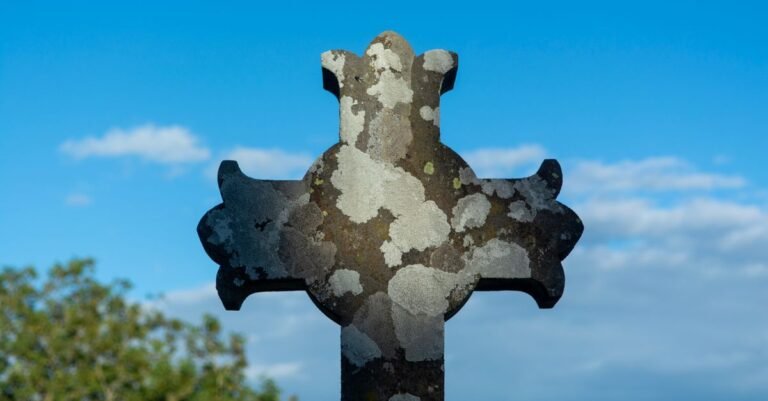 Pale crusty lichen spreading across the arms of a weathered stone cross against a blue sky