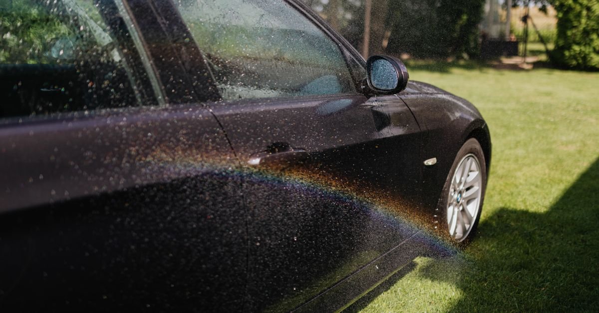 A dark car mid-wash with water sheeting off the paintwork and droplets in sunlight