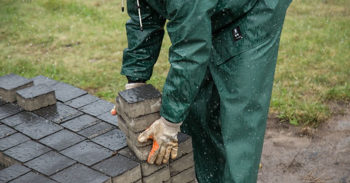 A worker in wet-weather gear setting paving stones during outdoor construction