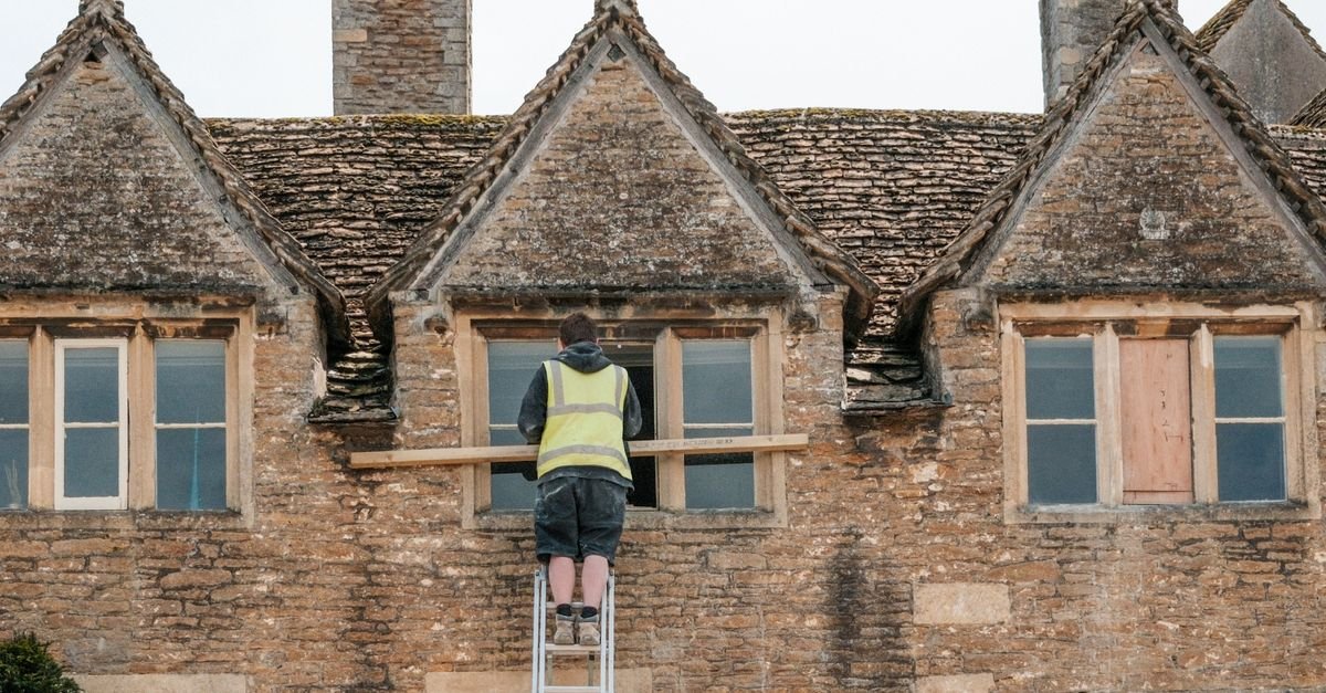 A worker on an extension ladder set against a stone building, working at the eaves