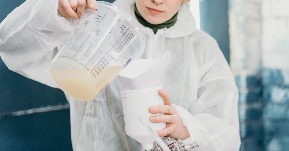 Gloved hands in protective coveralls pouring a measured amount of liquid cleaner into a mixing container