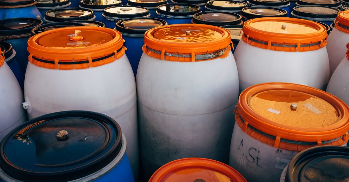 Blue and white plastic chemical barrels with orange lids stored in a shaded area