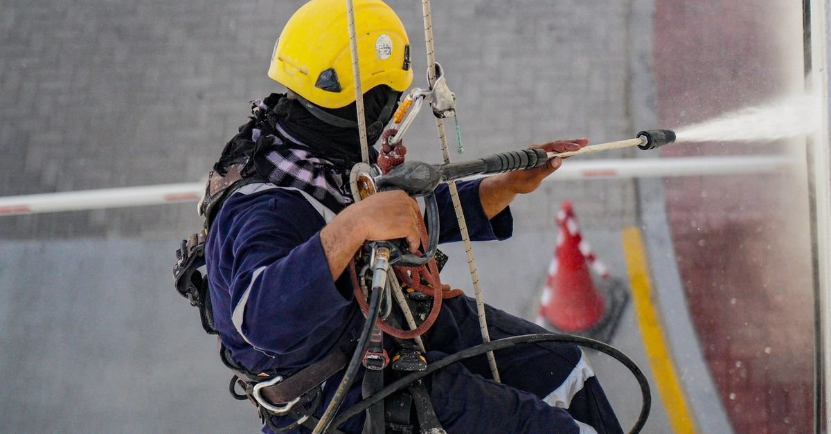 A worker in safety gear washing the face of a building from a raised position, spray angled downward across the wall