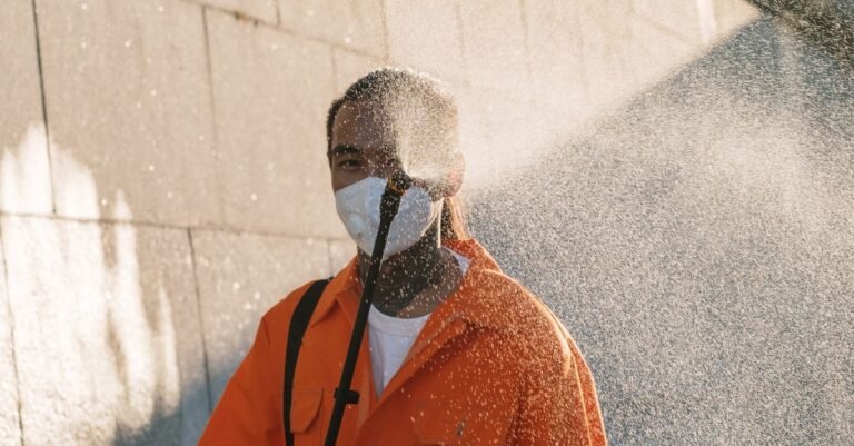 A person in a mask spraying water at an exterior wall with visible mist drifting across the surface
