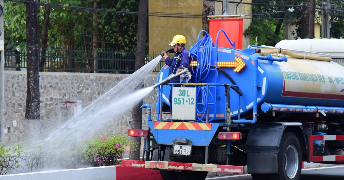 A street maintenance crew throwing a long low-pressure stream of water over roadside planting from a tanker