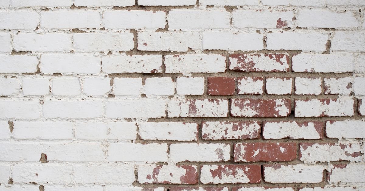 An old white-painted brick wall with the paint film peeling and blistering away from the masonry