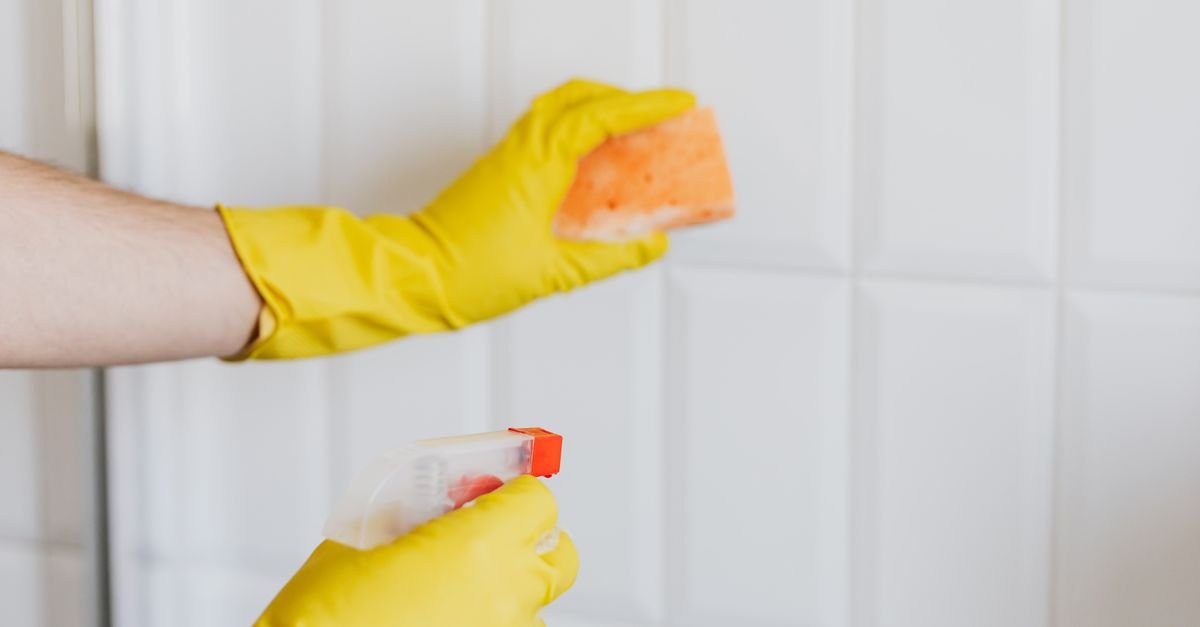 A person in yellow rubber gloves wiping a white tiled wall with a spray bottle and a soapy sponge