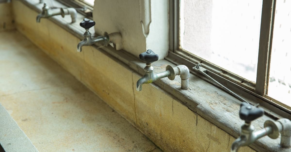 Row of aged metal taps over a dirty sink with pale mineral crust built up around the spouts