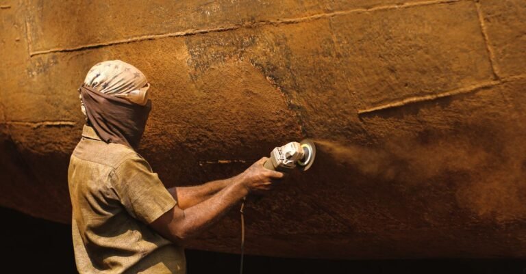 Worker in protective clothing grinding rust off a metal surface outdoors, throwing a shower of debris