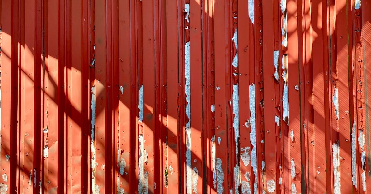 Close-up of a weathered red painted metal wall with the paint lifting and strong shadow across it