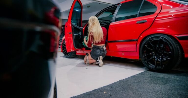 A woman kneeling in a garage to clean the interior carpet of a red hatchback by hand