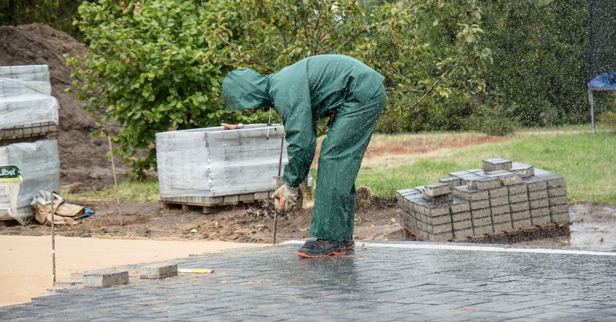 A worker in protective wet-weather gear bedding paving stones into place on a damp day