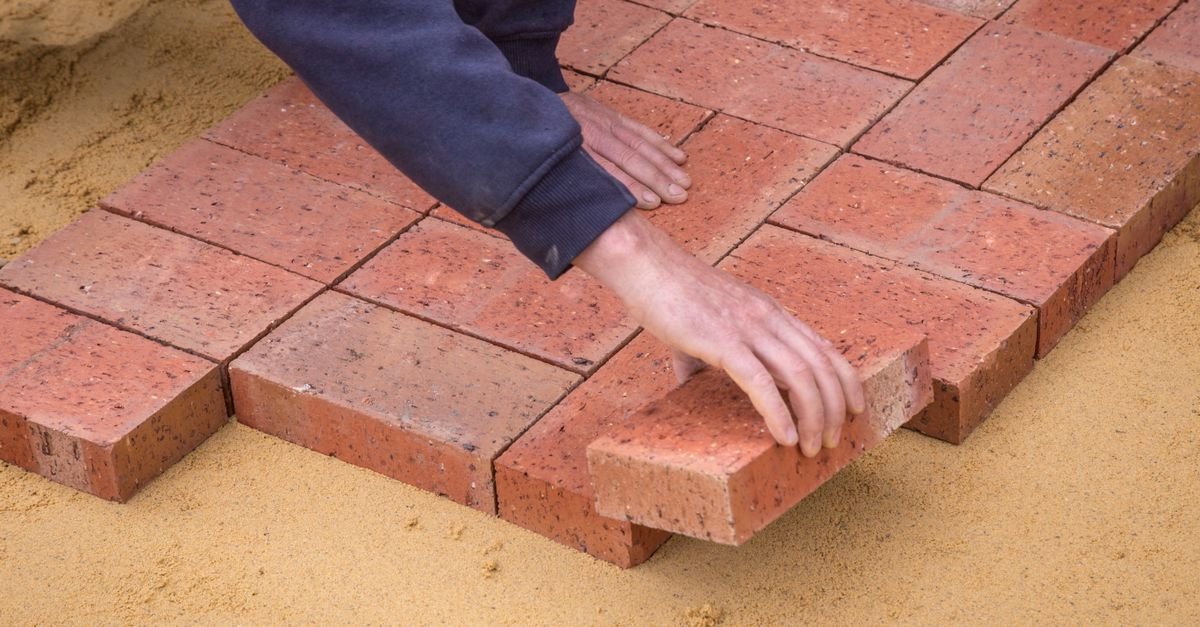 Overhead view of a worker in uniform laying brick pavers on a prepared sand bed