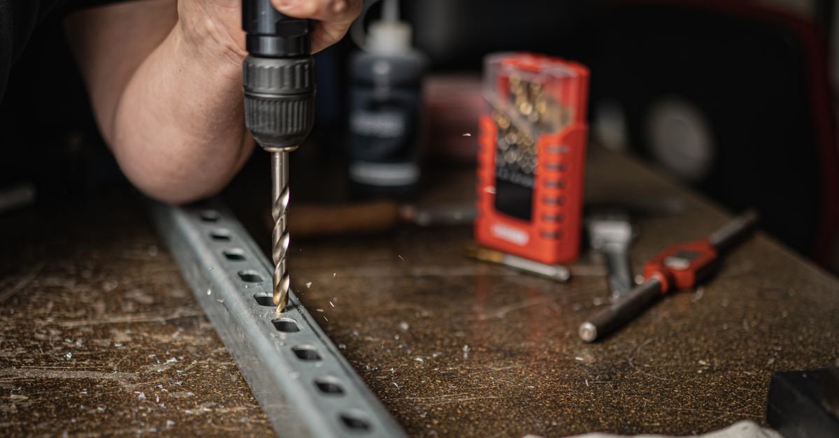 A hand holding a power drill, boring a hole through a sheet of metal indoors