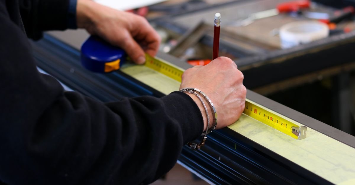 A worker measuring a length of aluminium stock with a tape measure on a workshop bench