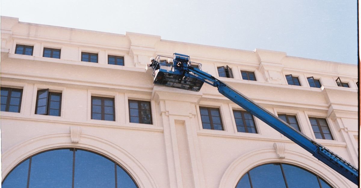 A worker cleaning the windows of a building from a blue crane on a clear day, the access method a pole is bought to replace